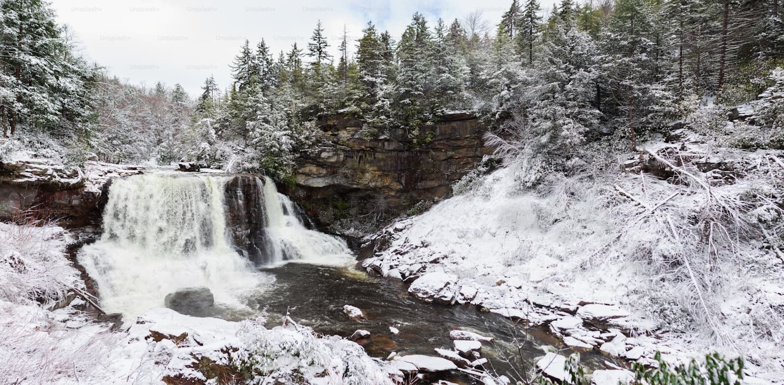 Snow-covered waterfall flowing through a winter forest with evergreen trees and rocky cliffs