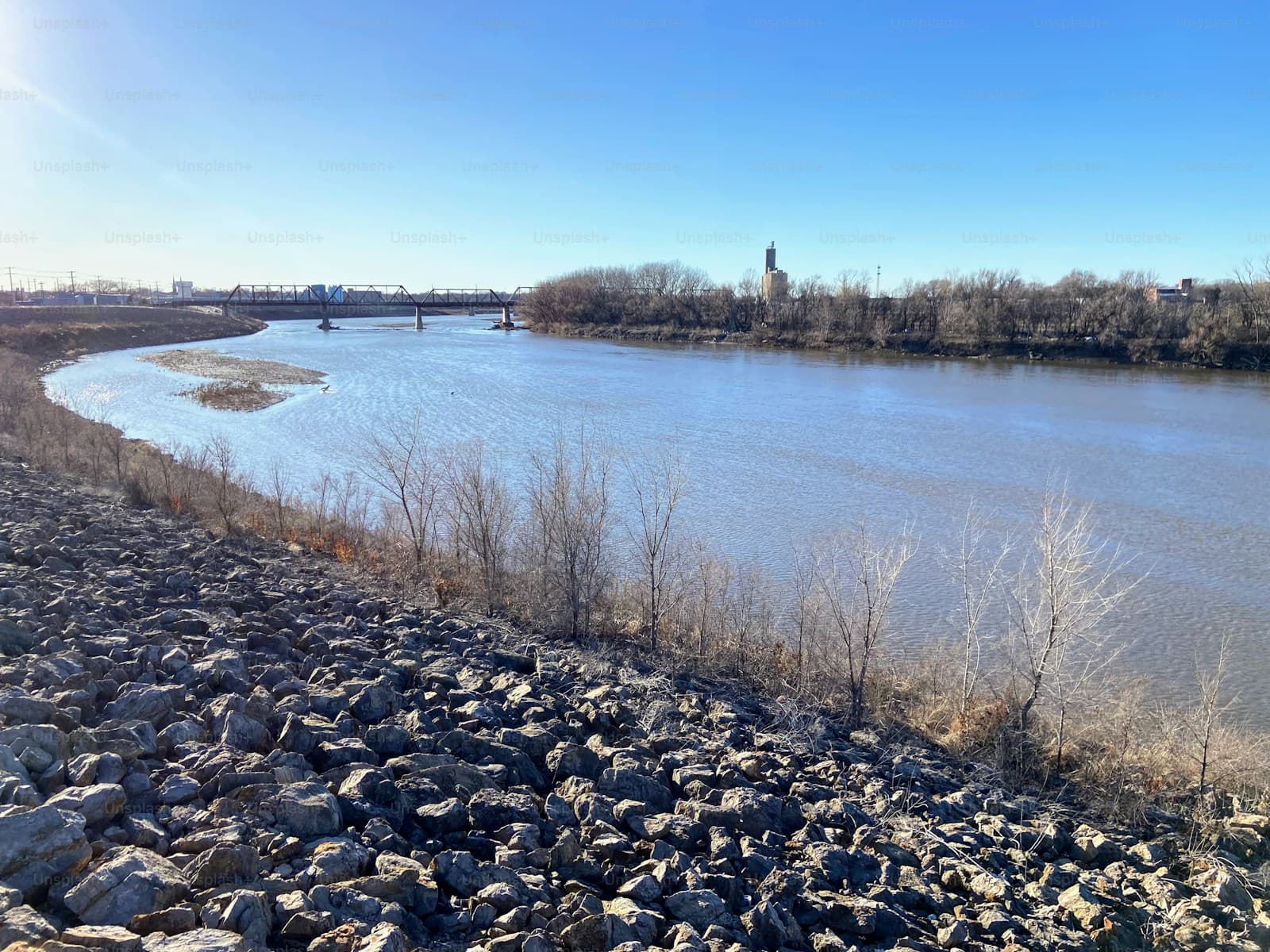 Wide river with rocky stone shoreline, bare trees on opposite bank, and bridge visible in distance under clear sky