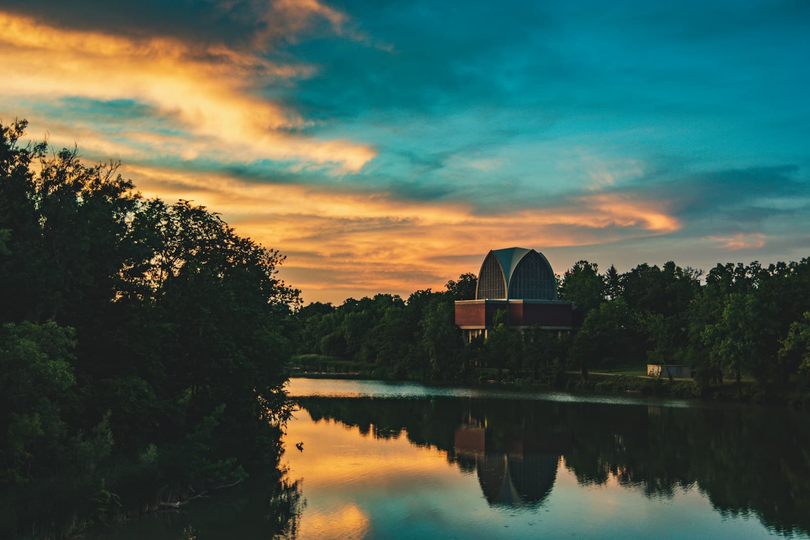 Sunset over a calm lake with orange clouds reflected in water, trees and barn silhouetted against colorful sky
