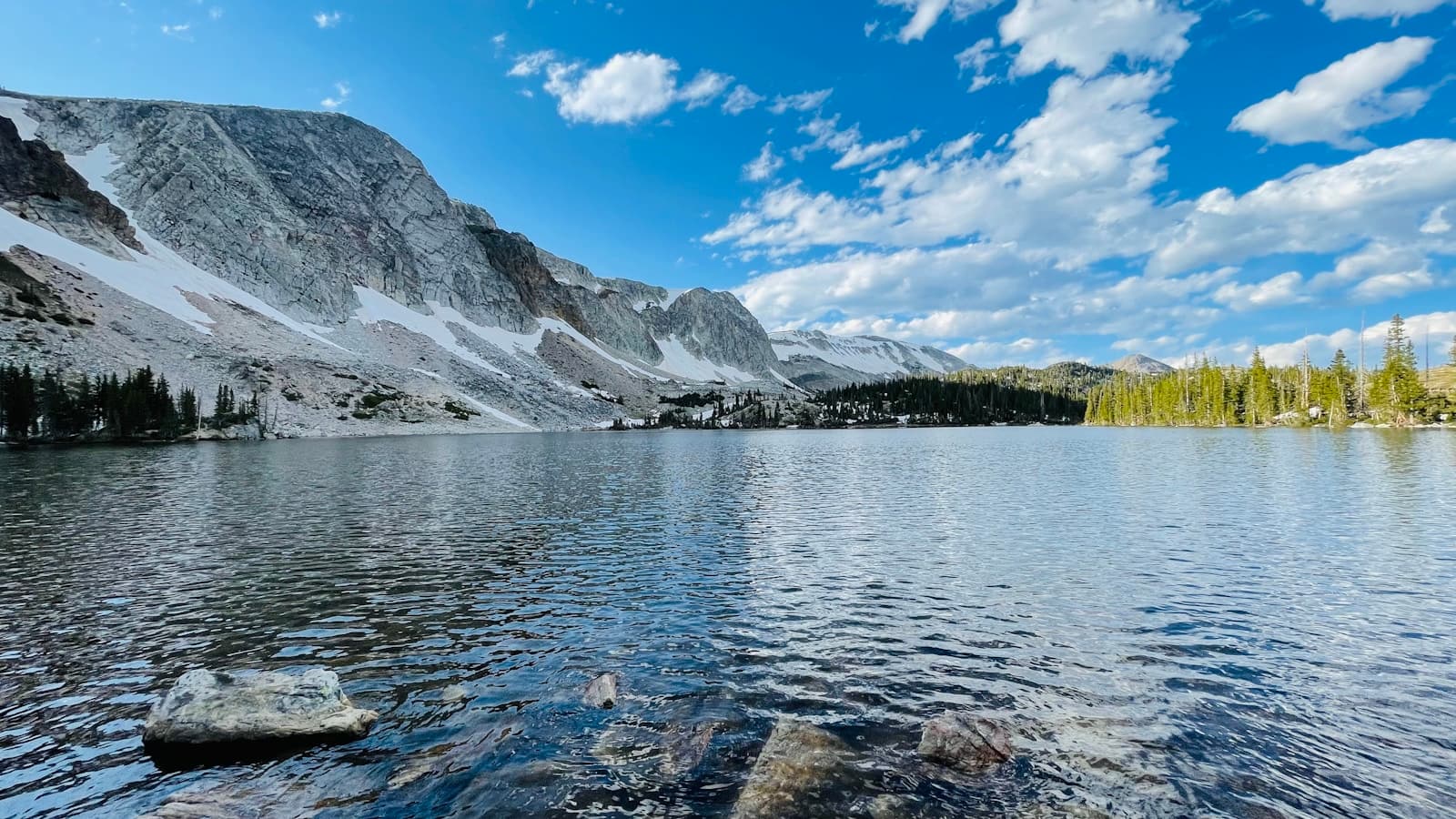 Alpine lake with snow-capped mountains, clear water, and evergreen trees under blue sky with clouds