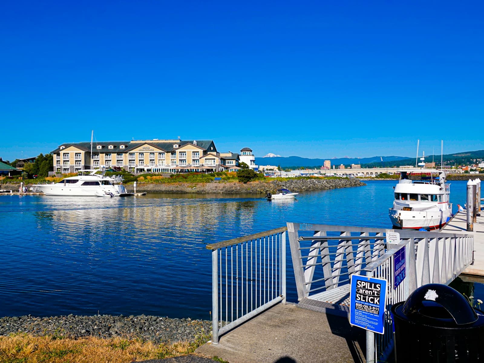 Harbor view with boats, waterfront buildings, and mountains under blue sky taken from pier with railing