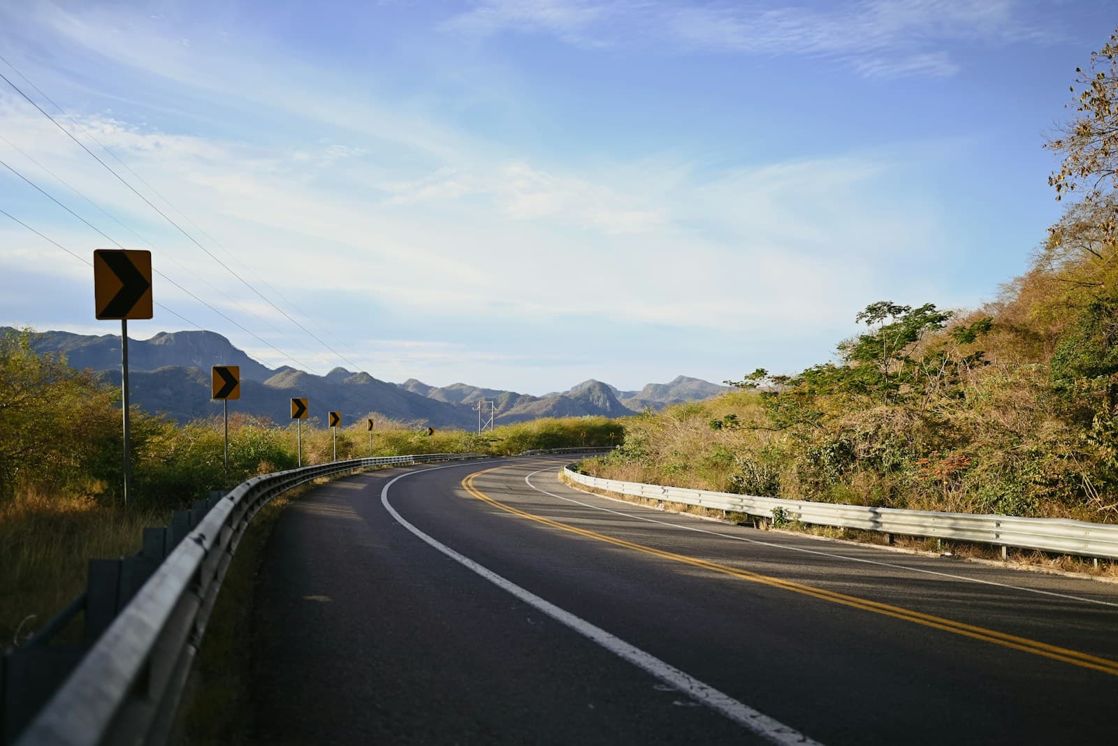 Curved highway through rolling hills with yellow warning signs along the guardrail