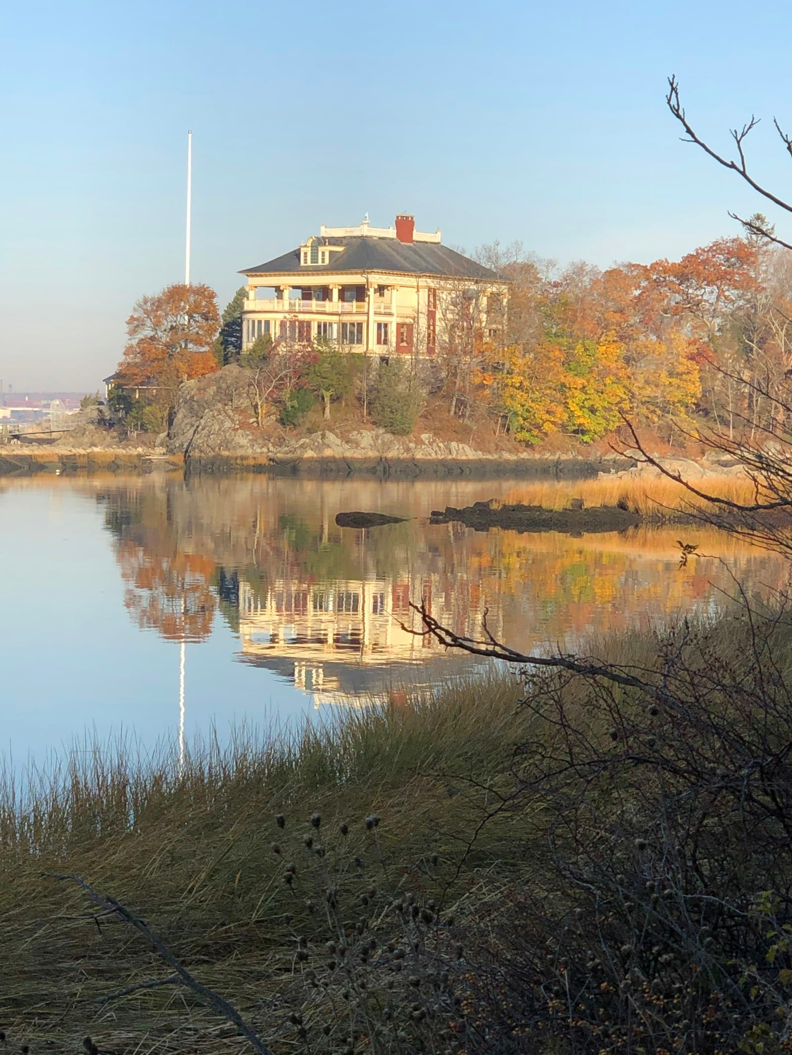 Colonial house with porch reflected in calm water, surrounded by autumn trees and tall flagpole
