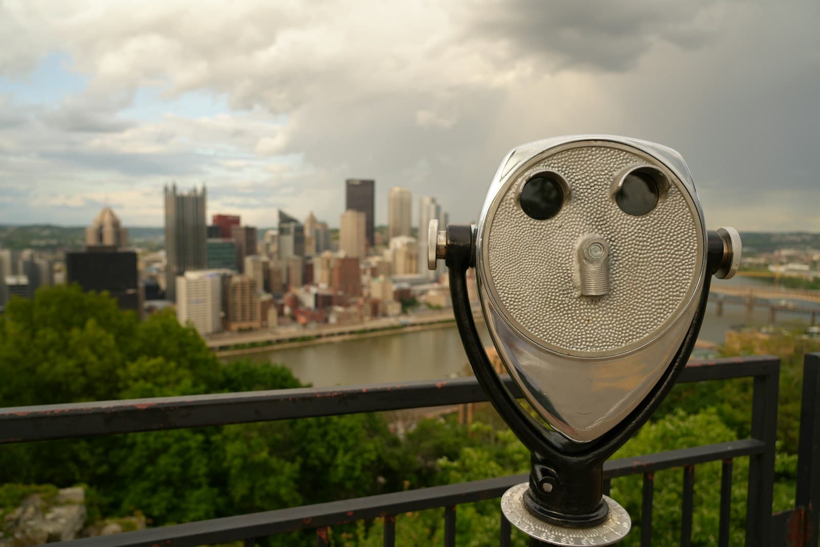 Coin-operated tourist telescope on viewing platform overlooking city skyline with buildings and river