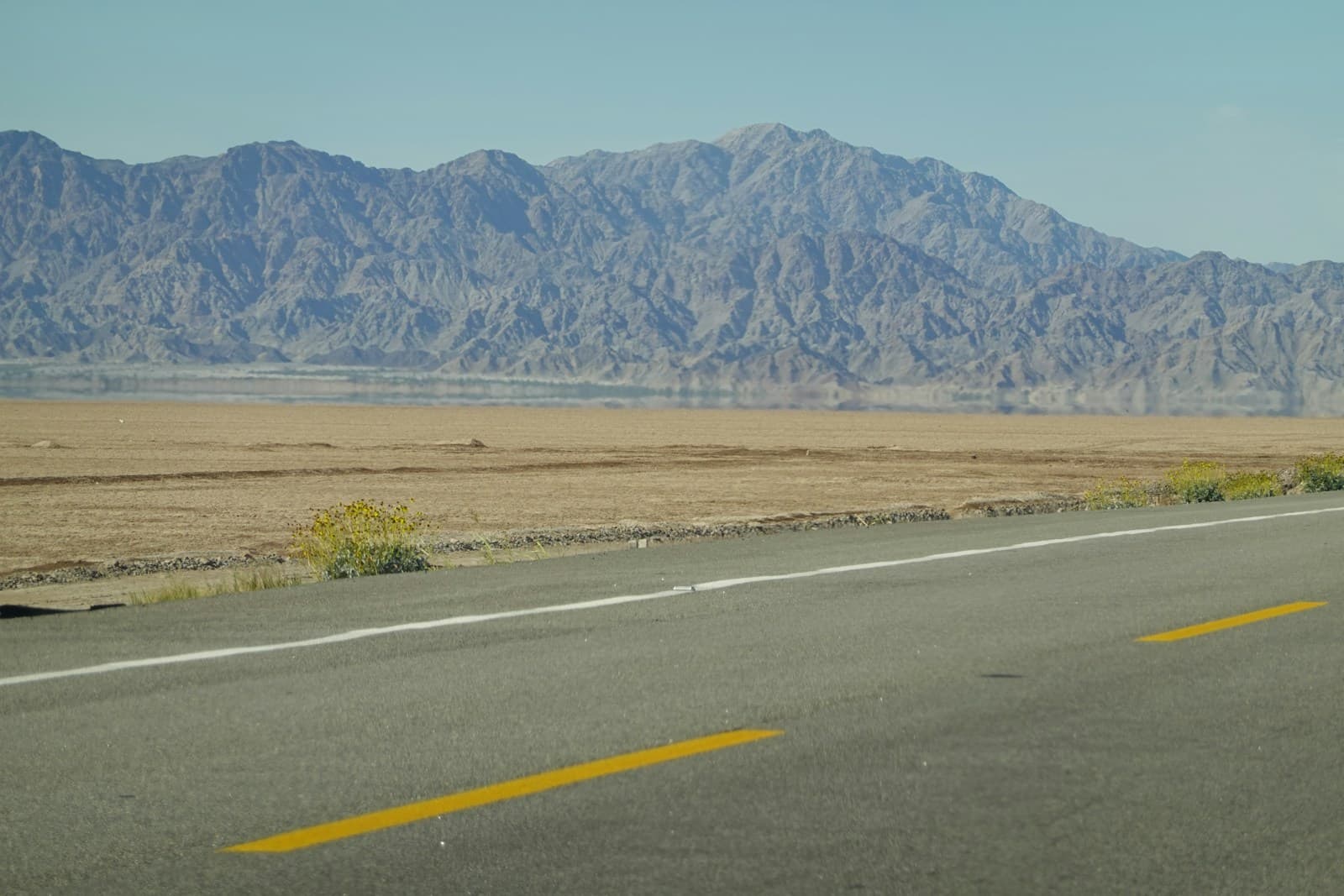 Desert highway with yellow lines leading toward distant mountains under blue sky in arid landscape