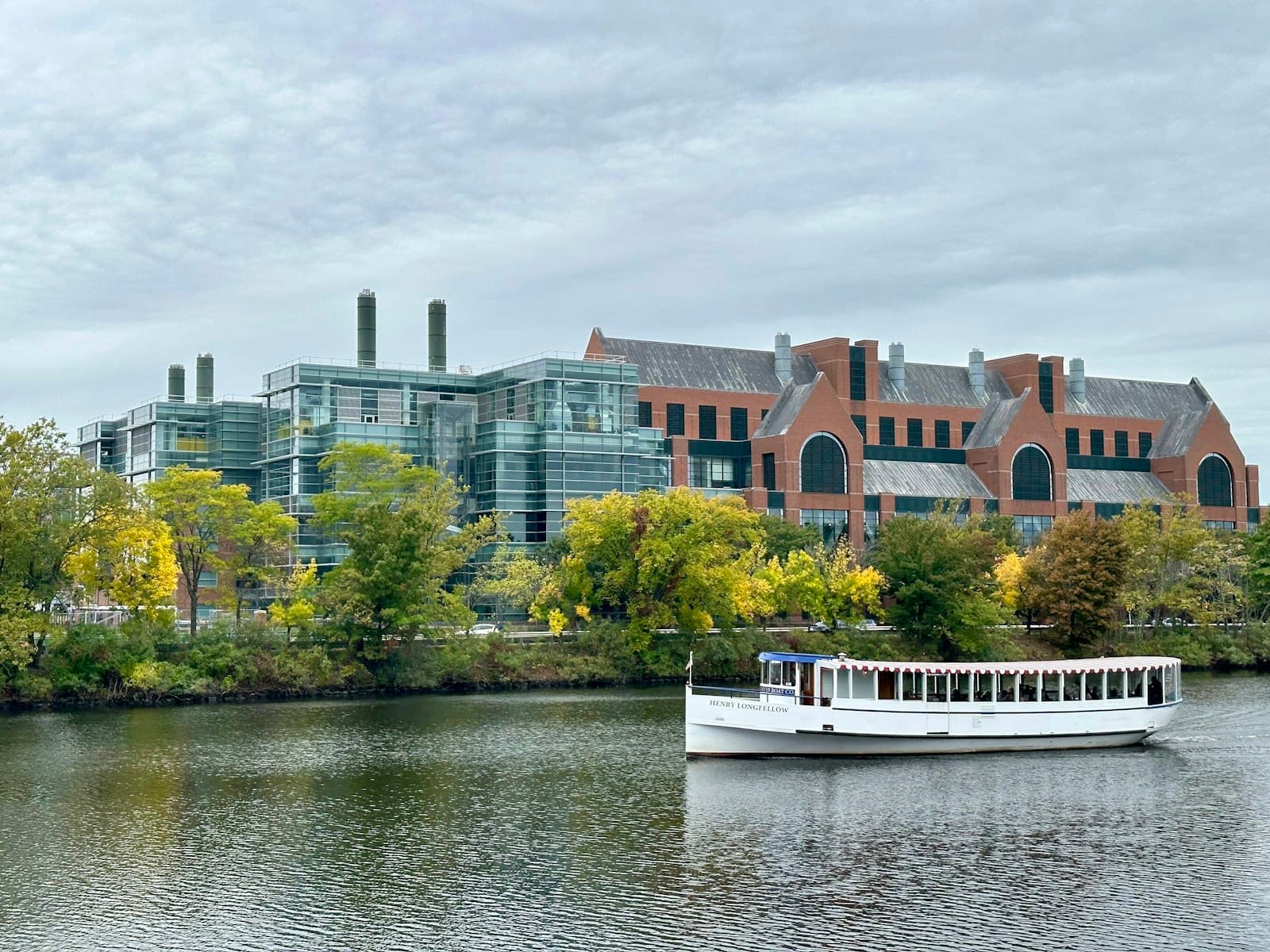White passenger boat on river with large brick and glass building complex and trees along shoreline