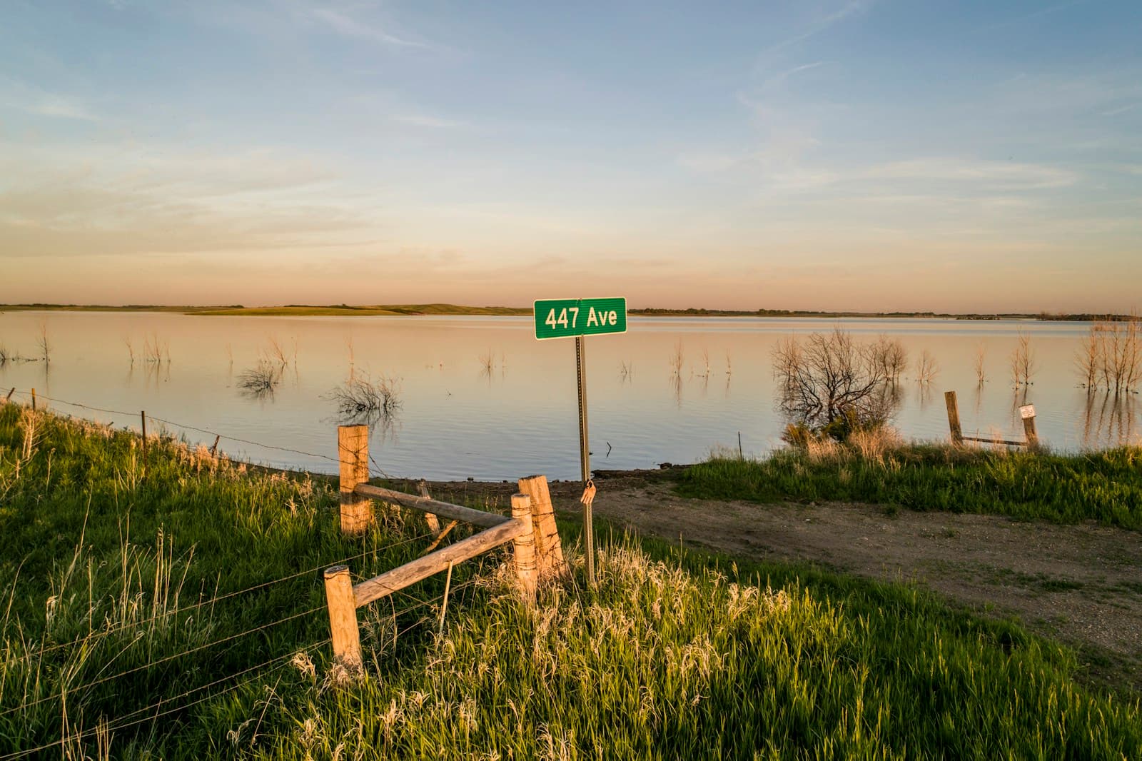 Rural flooded field with 447 Ave street sign, wooden fence, and dirt road under evening sky